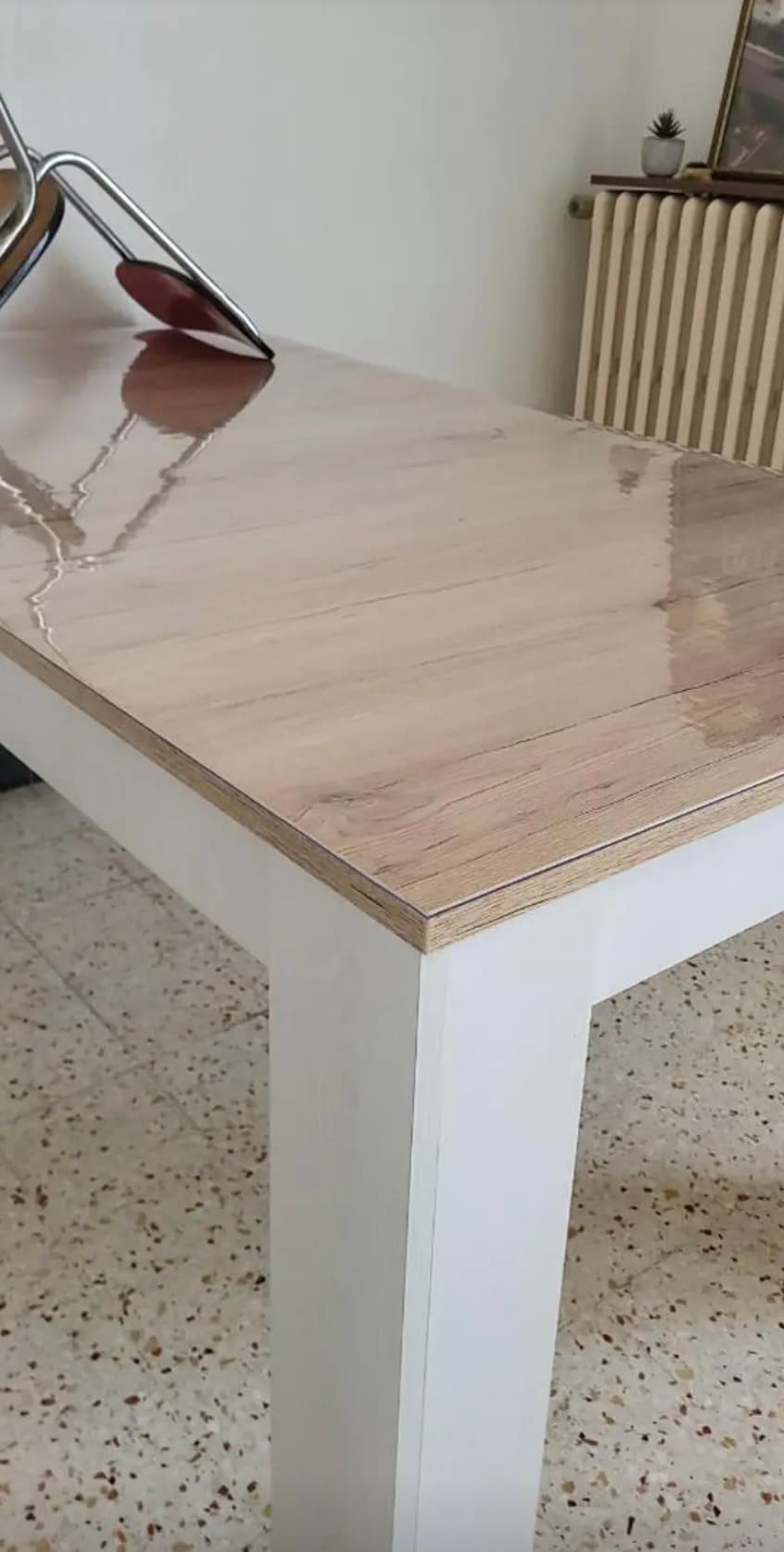 Jasny blat stołu na białych nogach z połyskiem maty; w tle żeliwny kaloryfer
